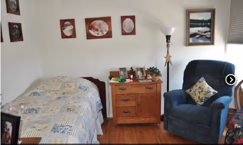A cozy bedroom with a single bed covered in a patterned quilt, a wooden nightstand with framed photos and small plants, a blue upholstered armchair with a decorative pillow, a floor lamp, and framed pictures on the walls.