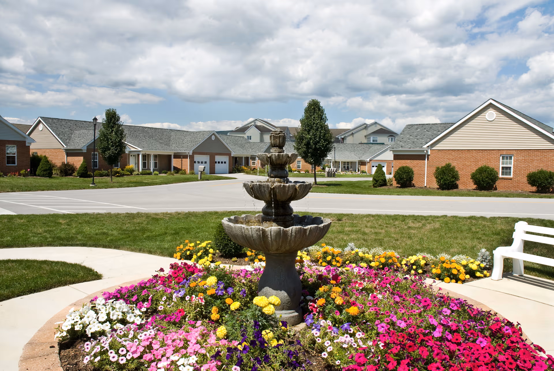 A tiered stone fountain surrounded by colorful flower beds in front of single-story brick residential buildings under a partly cloudy sky.