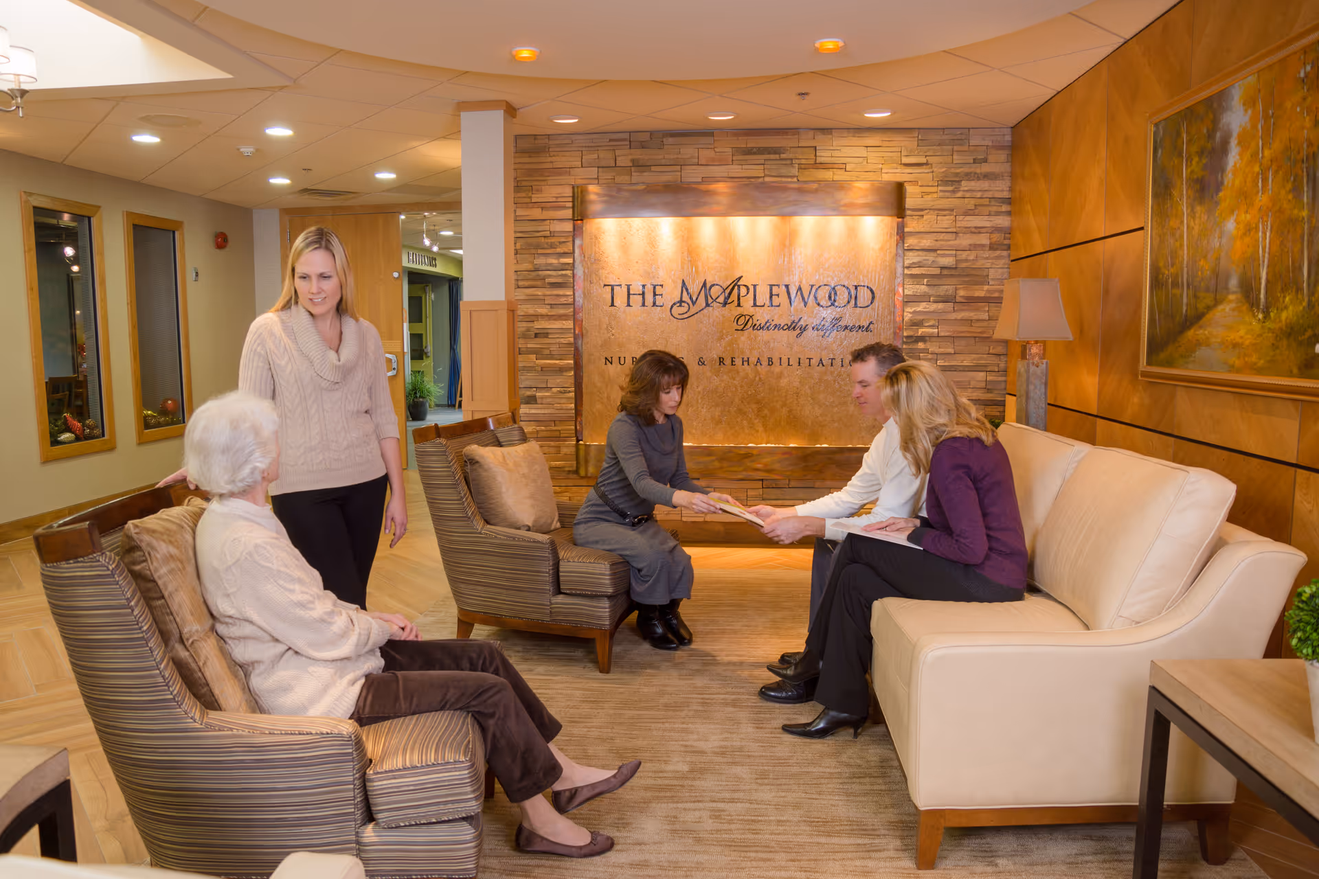A comfortable nursing home lobby with several adults seated and talking in front of a wall sign that reads 'The Maplewood'.
