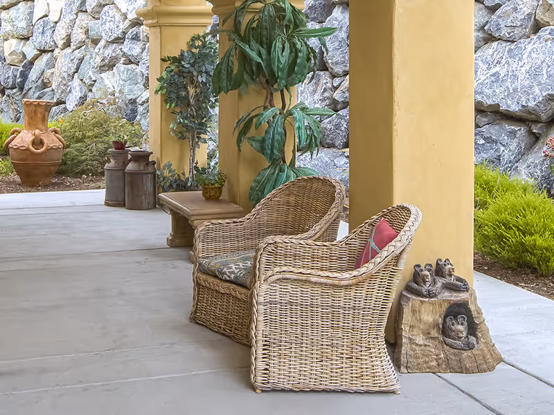 Outdoor patio area with two wicker chairs, one with a cushion and a red pillow, positioned near yellow columns. There are potted plants, decorative ceramic jars, and a carved wooden sculpture of bears on a tree stump. A stone wall and greenery are visible in the background.