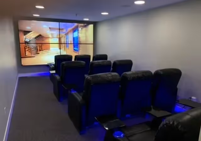 A small private theater room with two rows of black leather recliner chairs facing a large multi-panel screen on the wall. The room has dim lighting with blue accent lights under the chairs and a carpeted floor.