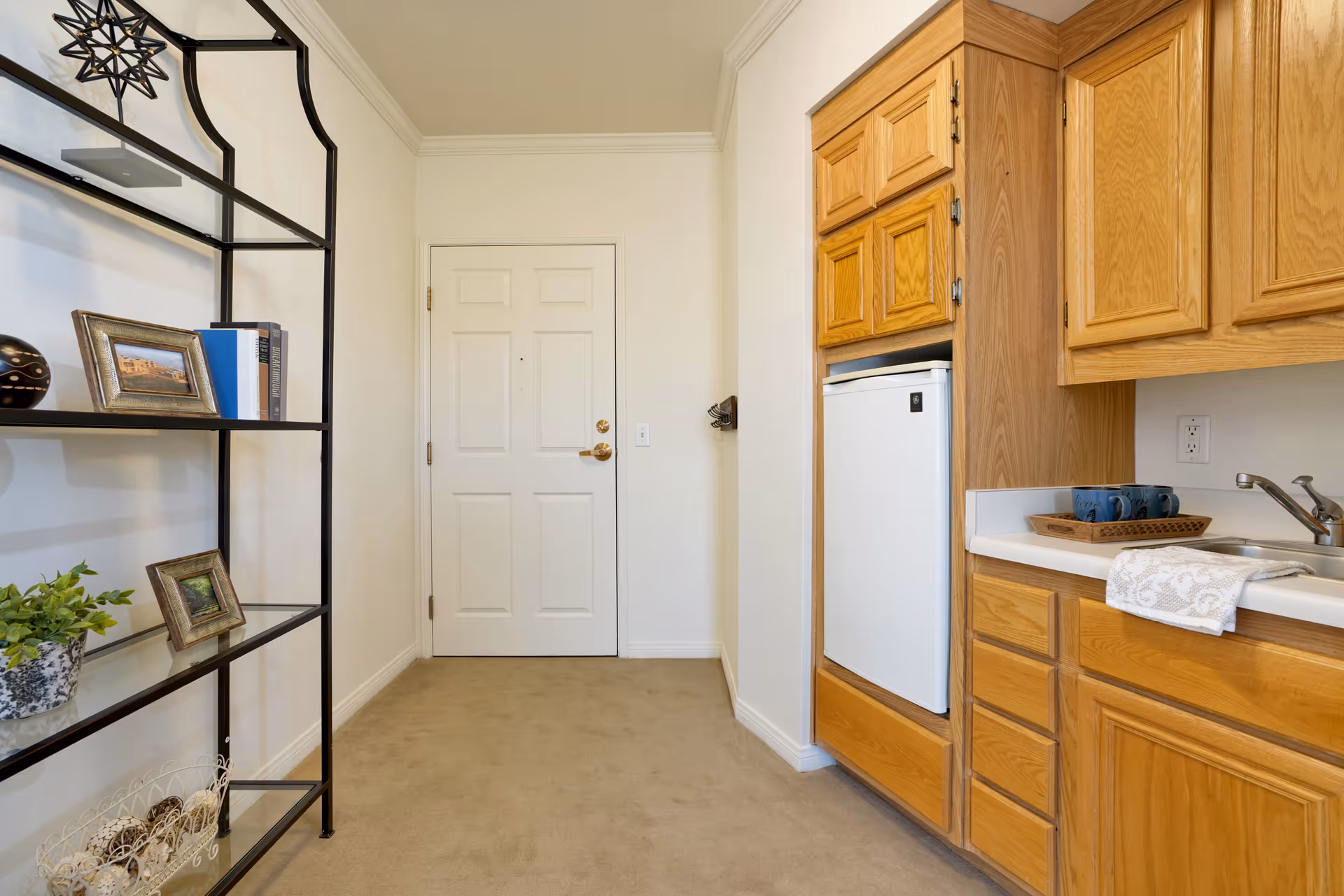Interior view of a small kitchenette area with wooden cabinets, a white mini refrigerator, a sink with a faucet, and a countertop with a tray holding two blue mugs and a folded towel. To the left, there is a black metal shelving unit with decorative items including framed photos, books, a plant, and a decorative bowl. A white door is visible at the end of the room.