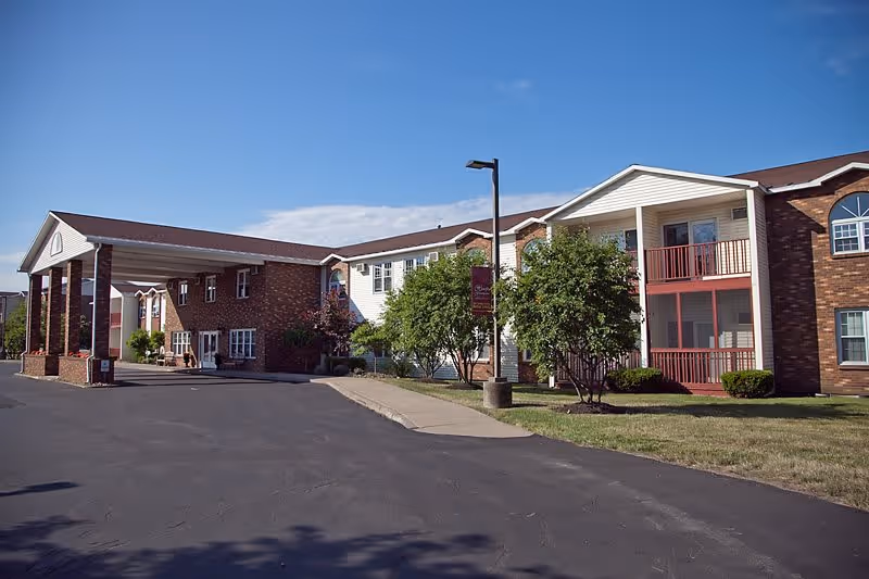 Exterior view of The Hearth at Greenpoint senior living facility showing a two-story building with brick and white siding, a covered entrance with columns, trees, and a paved driveway under a clear blue sky.