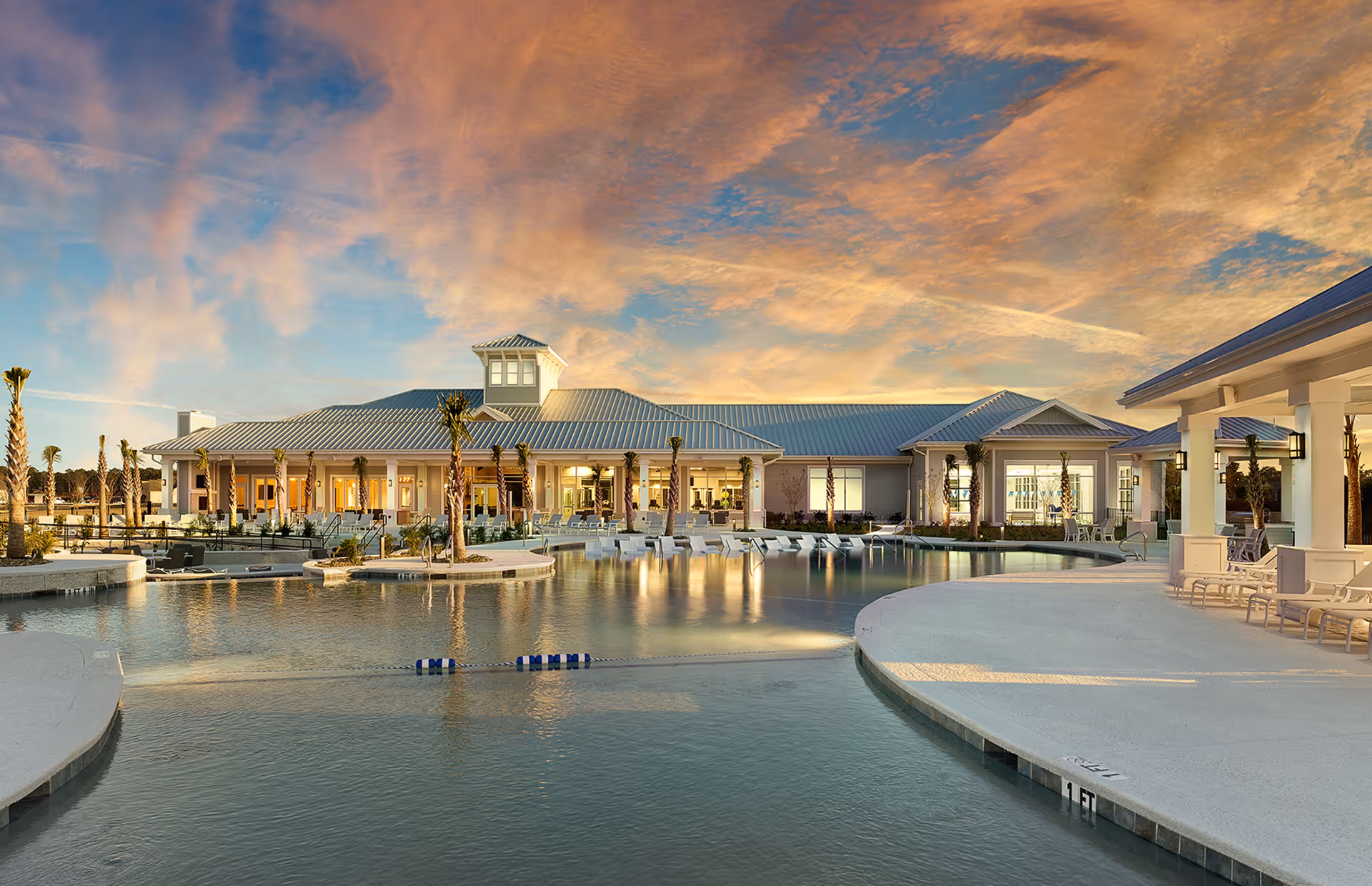 A large outdoor swimming pool with lounge chairs around it, set in front of a single-story building with a metal roof and large windows during sunset with a colorful sky.