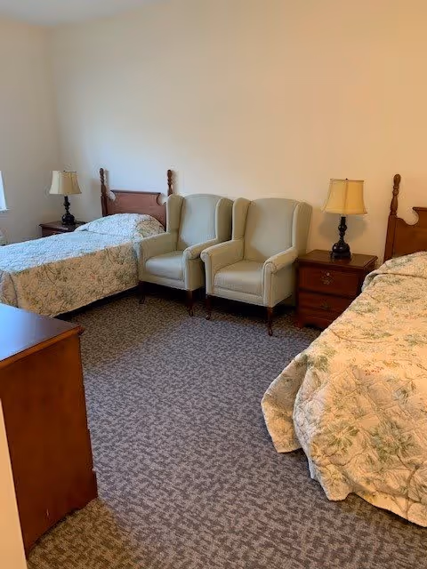 A bedroom with two twin beds, each with a floral bedspread. Between the beds are two beige upholstered armchairs and a wooden nightstand with a lamp. Another wooden piece of furniture is partially visible in the foreground. The room has beige walls and carpeted flooring.