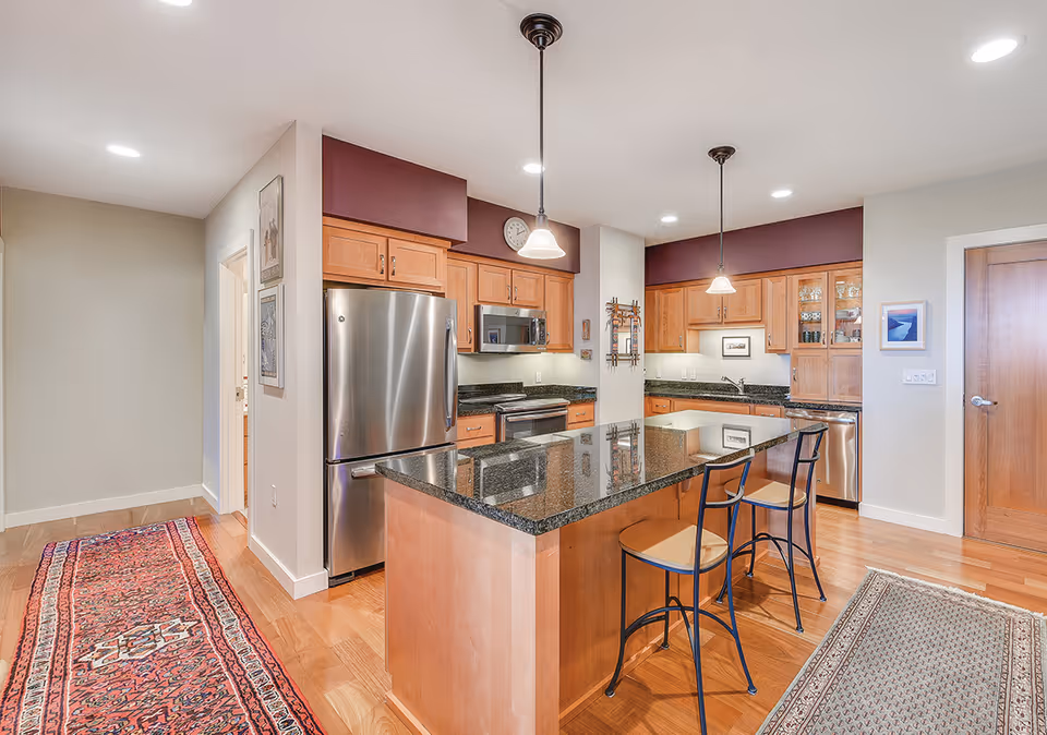 Modern kitchen with a large granite island, wooden cabinets, stainless steel appliances, and two bar stools.