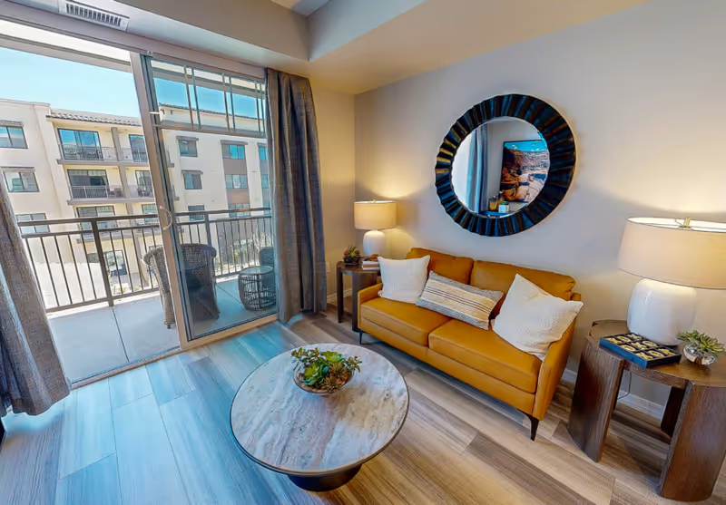 Bright living room with a mustard-yellow sofa, round mirror, coffee table, side tables with lamps, and sliding glass doors opening to a balcony.