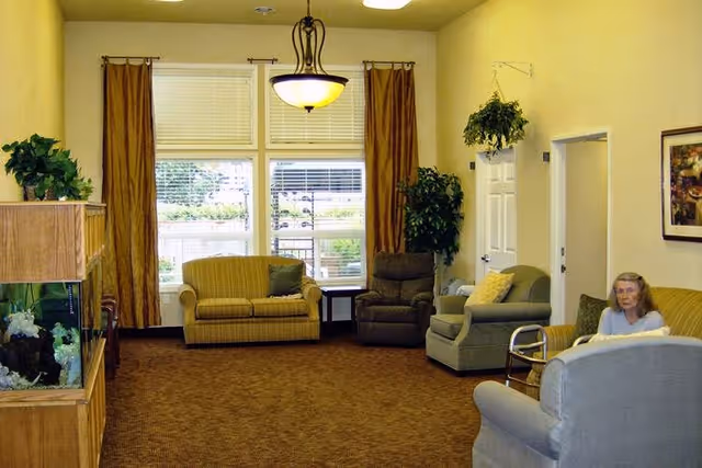A cozy living room in a memory care facility with beige walls and carpet. The room features a yellow loveseat, a gray recliner, a green armchair, and another gray armchair. There is a large window with brown curtains letting in natural light. A hanging plant is near a white door, and a framed picture is on the wall. A woman is seated on the yellow loveseat, and a fish tank is visible on the left side of the image.