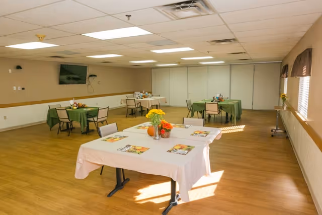 A spacious dining room with several tables covered in white and green tablecloths. Each table has chairs around it and is decorated with small flower arrangements and placemats. The room has wood flooring, beige walls, a wall-mounted TV, and large windows with brown valances letting in natural light.