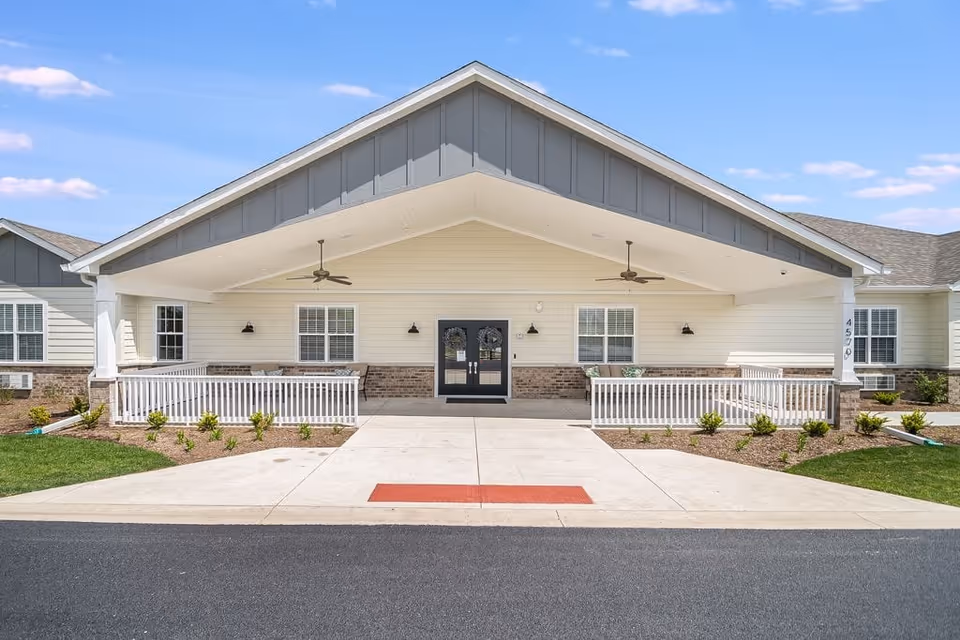 Front exterior view of The Heathers Senior Homes building with a covered entrance, double doors, white railings, ceiling fans, and landscaped greenery under a blue sky.