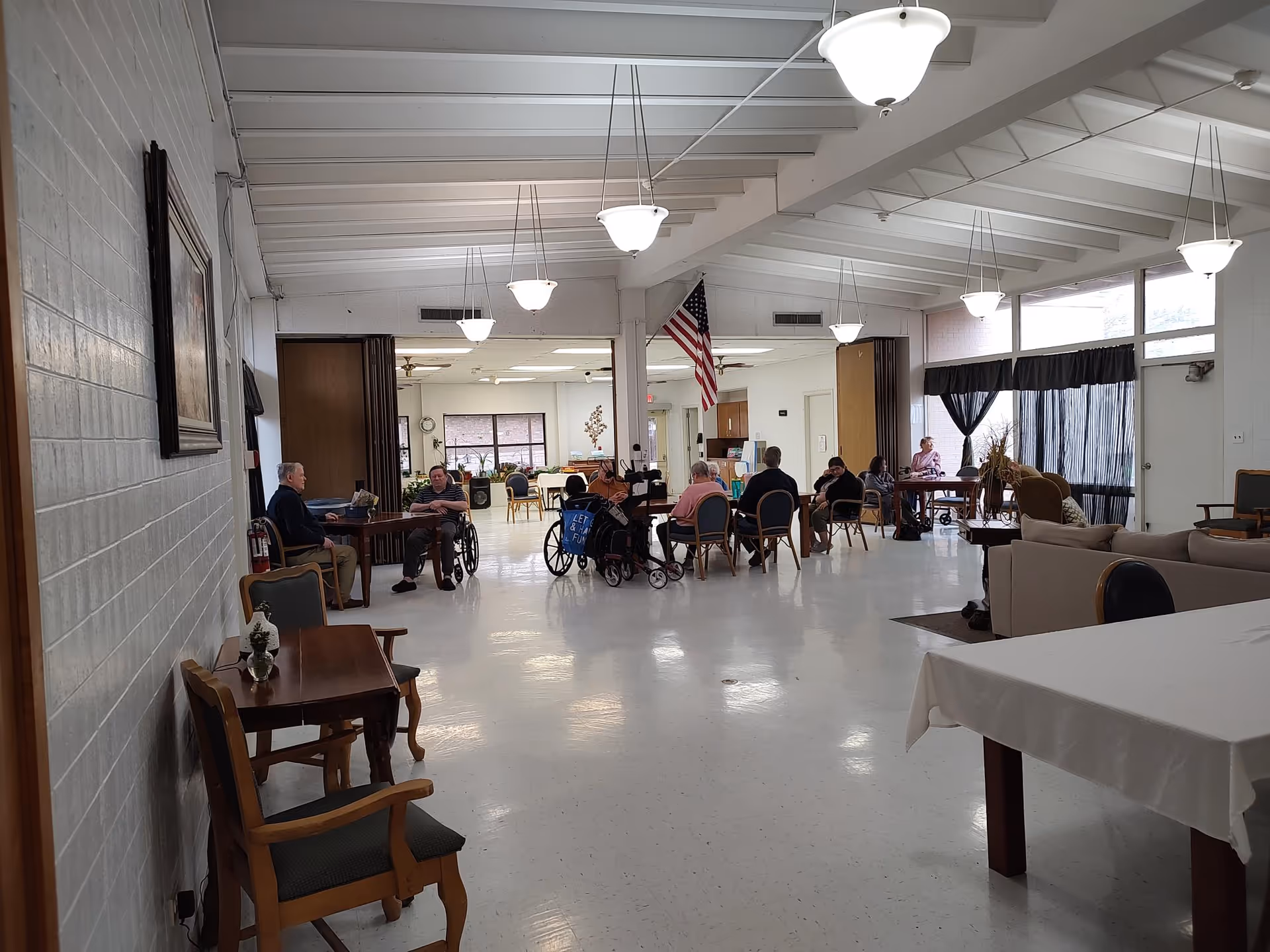 Spacious common dining/activity room in an assisted living facility with residents seated at tables and in wheelchairs under pendant lights.