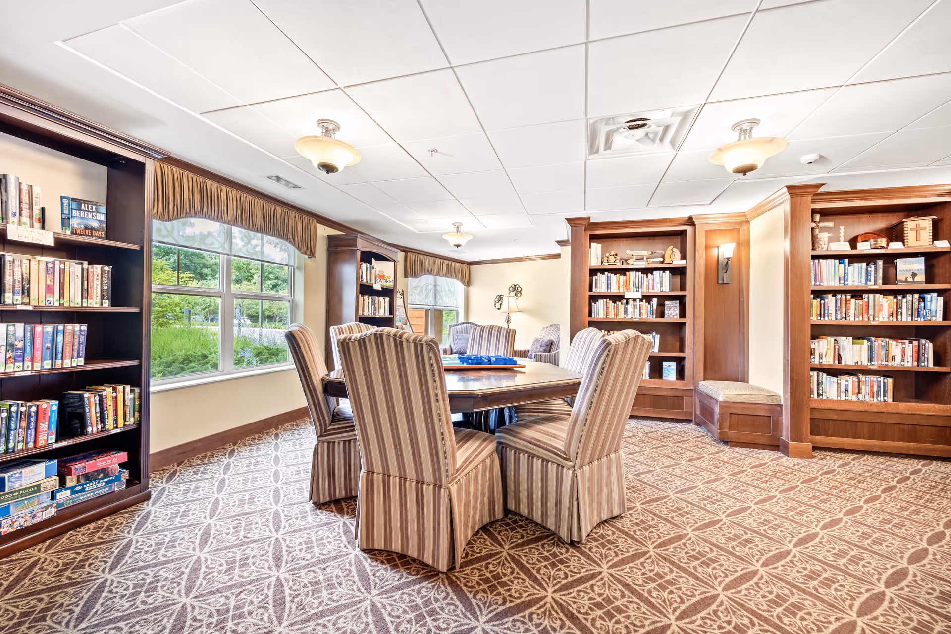 Bright senior living common room/library with a round table, striped chairs, bookshelves and large windows.