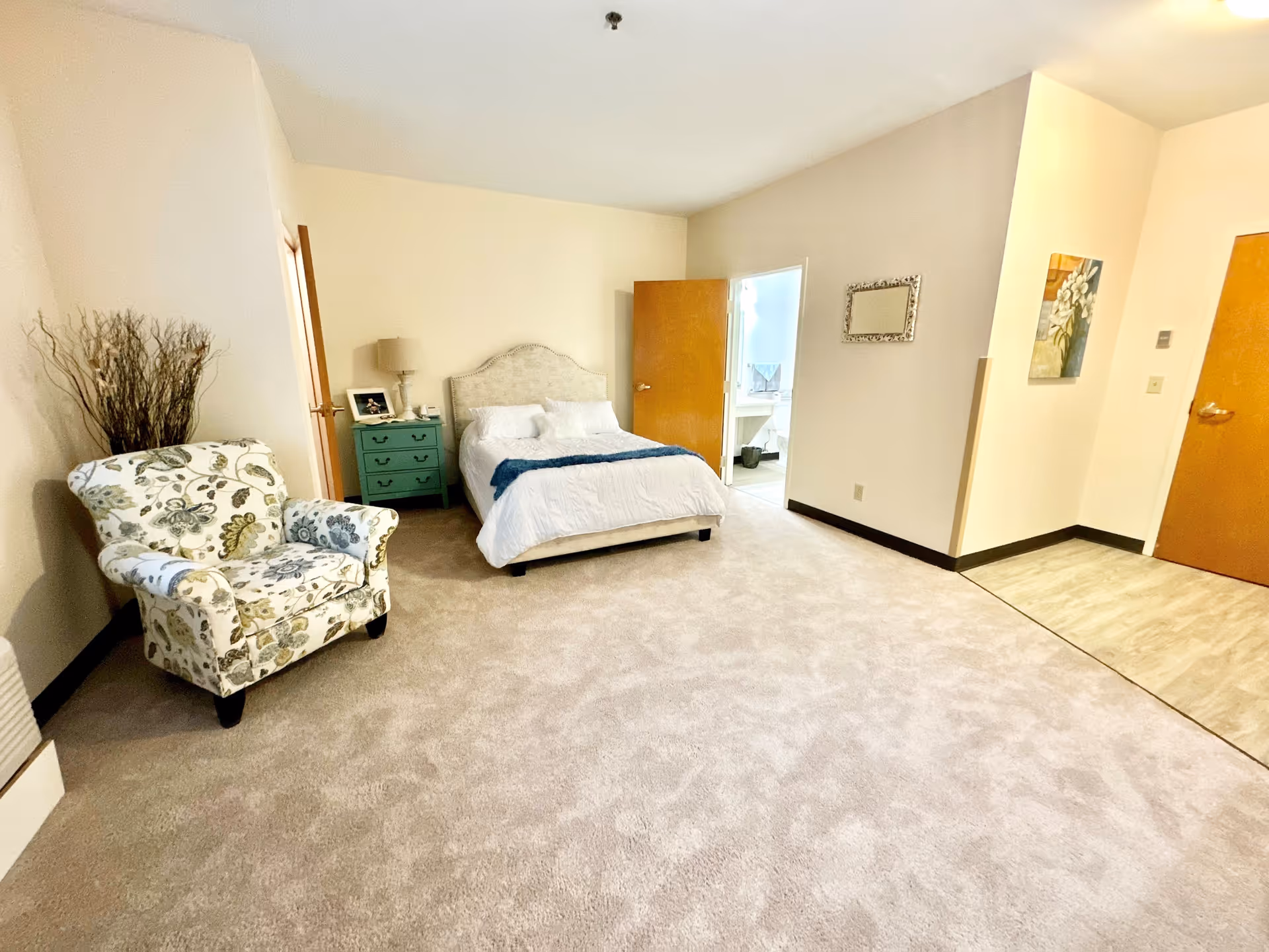 A spacious bedroom in a senior living community featuring a bed with white bedding and a blue throw, a green nightstand with a lamp and picture frame, a floral upholstered armchair, beige carpet, and an open door leading to a bathroom with a sink and mirror.