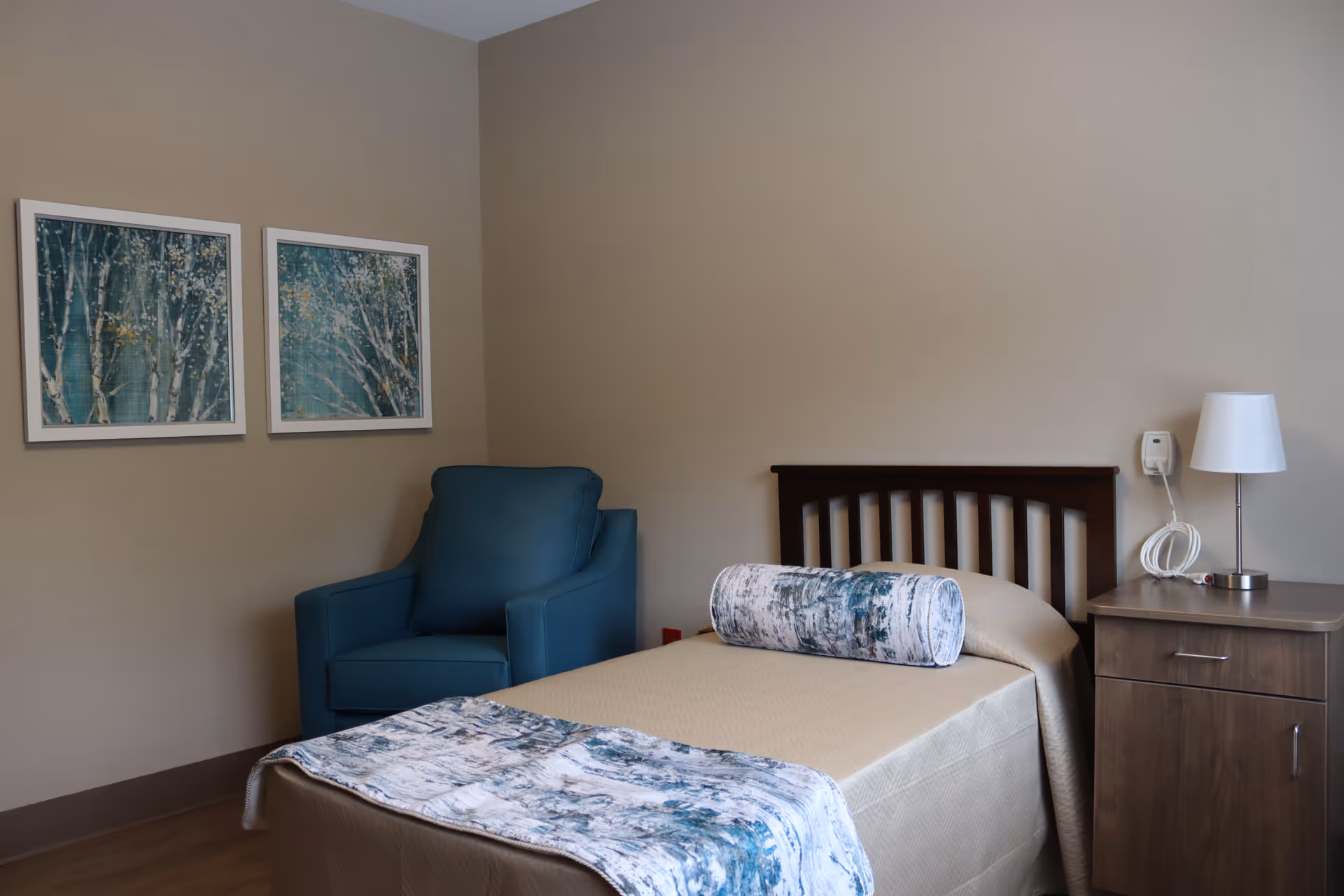 A neatly made single bed with a beige bedspread and a decorative cylindrical pillow in a room. Next to the bed is a wooden nightstand with a white lamp and a call button device. A blue armchair is positioned in the corner near two framed pictures of trees on the beige wall.