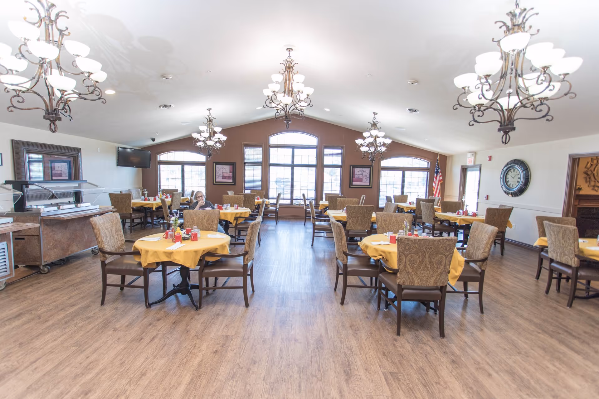 Spacious dining room with round tables draped in yellow tablecloths, upholstered chairs, chandeliers, and large windows.