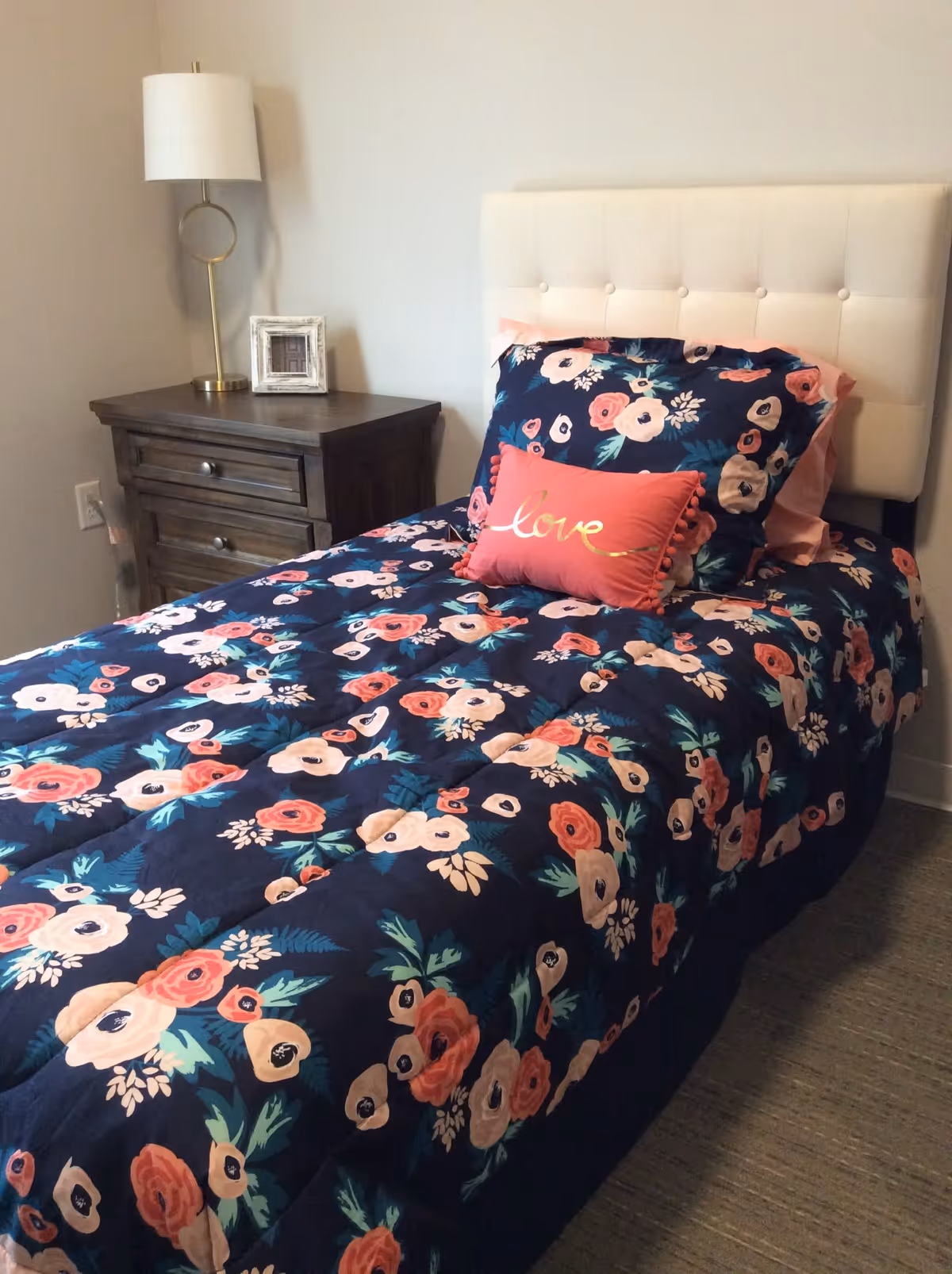 A single bed with a navy floral comforter and a pink 'love' pillow next to a wooden nightstand and lamp in a bedroom.