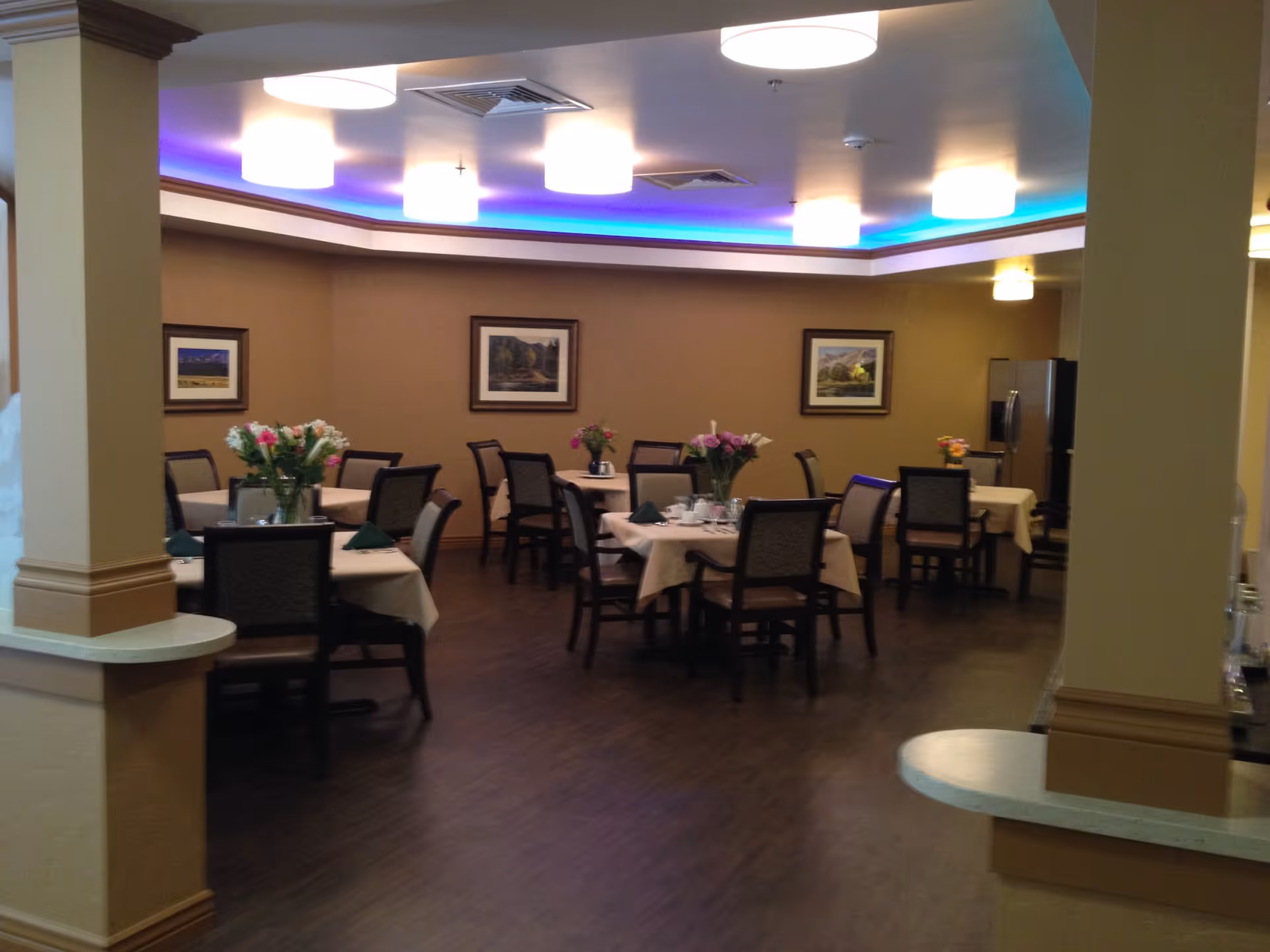 Interior view of a senior living community dining room with several tables covered in beige tablecloths, each set with chairs and floral centerpieces. The room has warm brown walls, framed landscape paintings, and a ceiling with recessed lighting and blue accent lights.