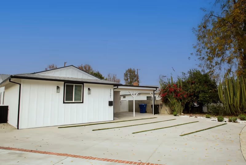 White, single-story building with a covered entrance, wide concrete driveway, and desert landscaping.