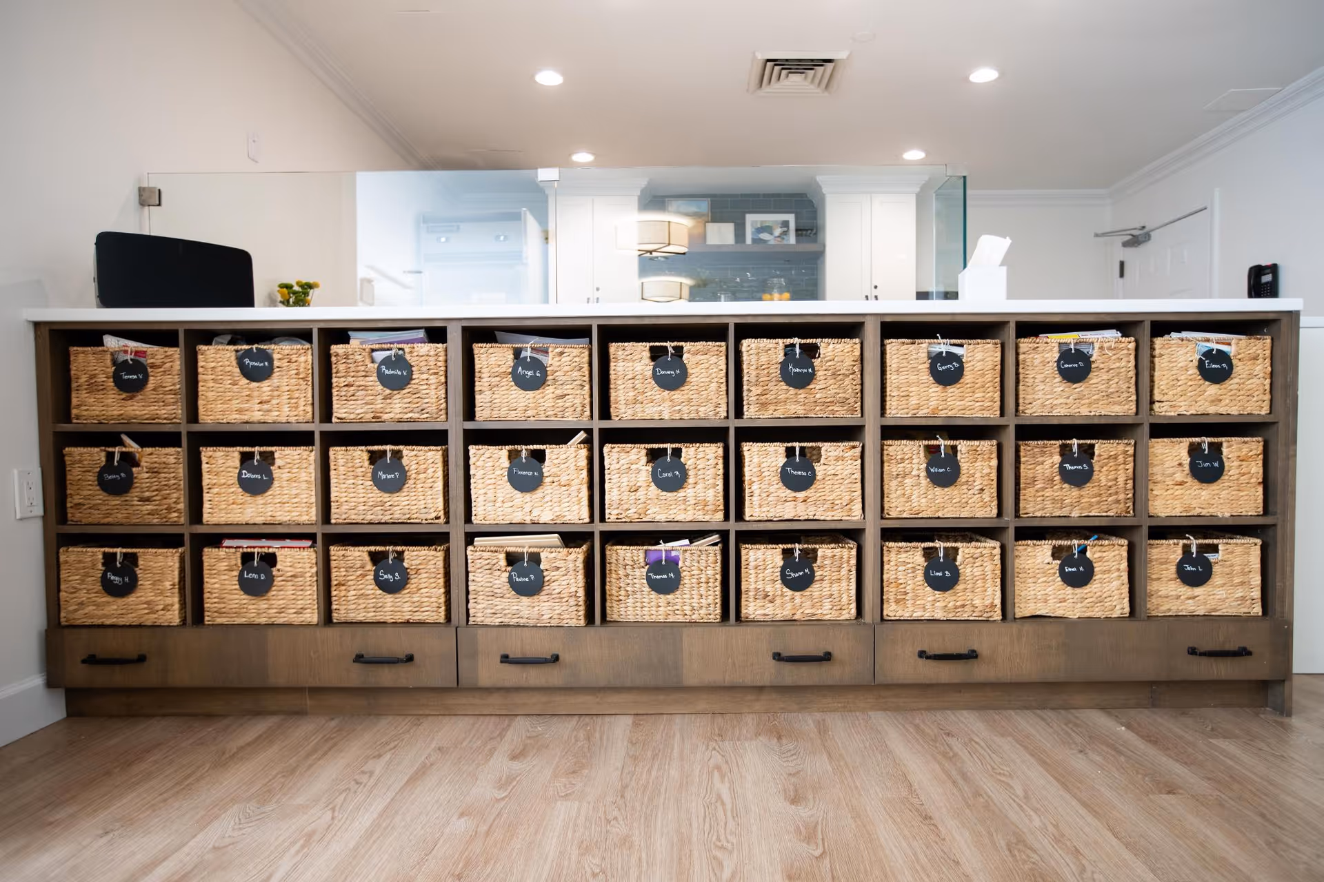 A wooden storage unit with multiple woven baskets, each labeled with a name tag, in a well-lit room with light wood flooring and white walls. The top of the unit has a white countertop with a glass partition behind it, and the background shows a door and some kitchen cabinets.