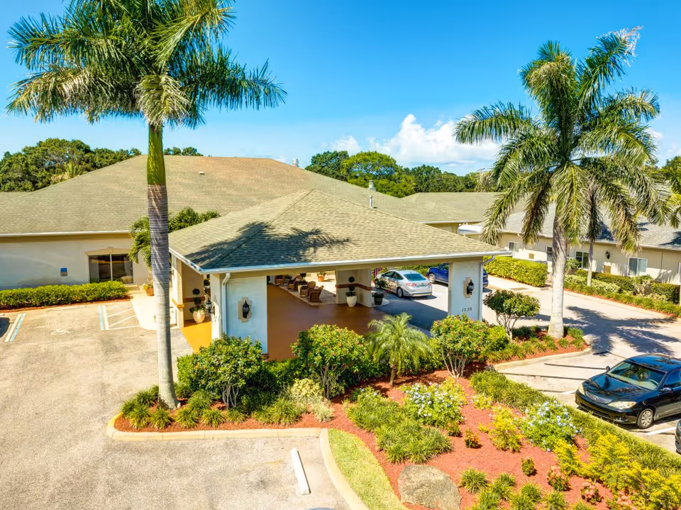 Exterior view of a senior living facility entrance with a covered drop-off area, surrounded by palm trees, landscaped bushes, and parked cars under a clear blue sky.