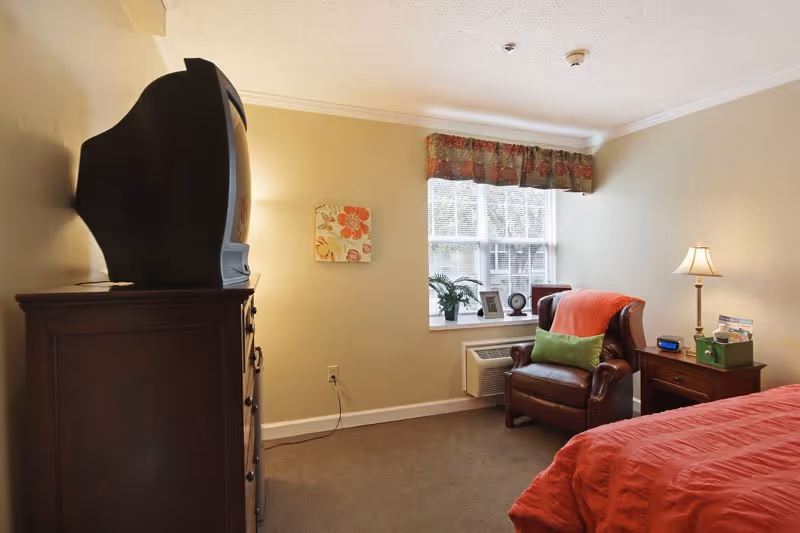A cozy bedroom with a large window covered by a floral valance. There is a brown leather armchair with a green pillow and an orange throw blanket next to a wooden nightstand with a lamp, clock, and some items. A wooden dresser with an older-style CRT television on top is positioned against the opposite wall. The bed has a red comforter, and the room has beige walls and carpeted floor.
