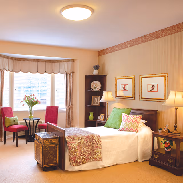 A cozy bedroom with a single bed adorned with colorful pillows and a patterned throw. There are two framed pictures on the wall above the bed. Beside the bed is a wooden nightstand with a lamp and small decorative items. A wooden shelving unit with decorative plates and a framed photo is in the corner. Near a large window with beige curtains, there are two red chairs with green cushions and a small round table with a vase of flowers. The room has warm lighting and a beige carpet.