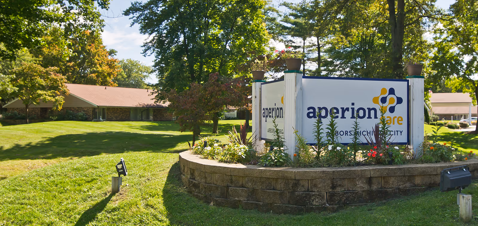 Landscaped entrance sign for Aperion Care surrounded by flowers and trees with single-story buildings in the background.