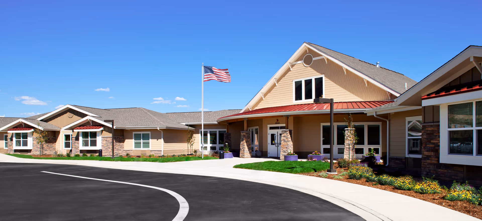 Exterior view of a single-story senior living facility building with beige siding, stone accents, and a red metal roof. There is a curved driveway in front, a flagpole with an American flag, and landscaped areas with flowers and small trees under a clear blue sky.