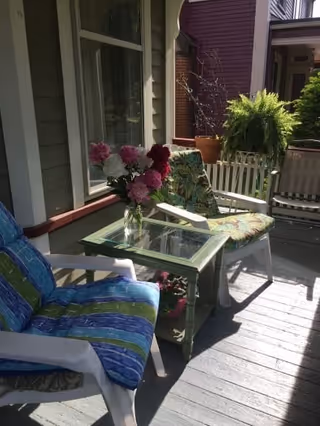 A sunlit porch area with cushioned chairs featuring colorful striped and floral patterns, a small green table with a glass top holding a vase of pink and white flowers, and a large potted fern in the background.