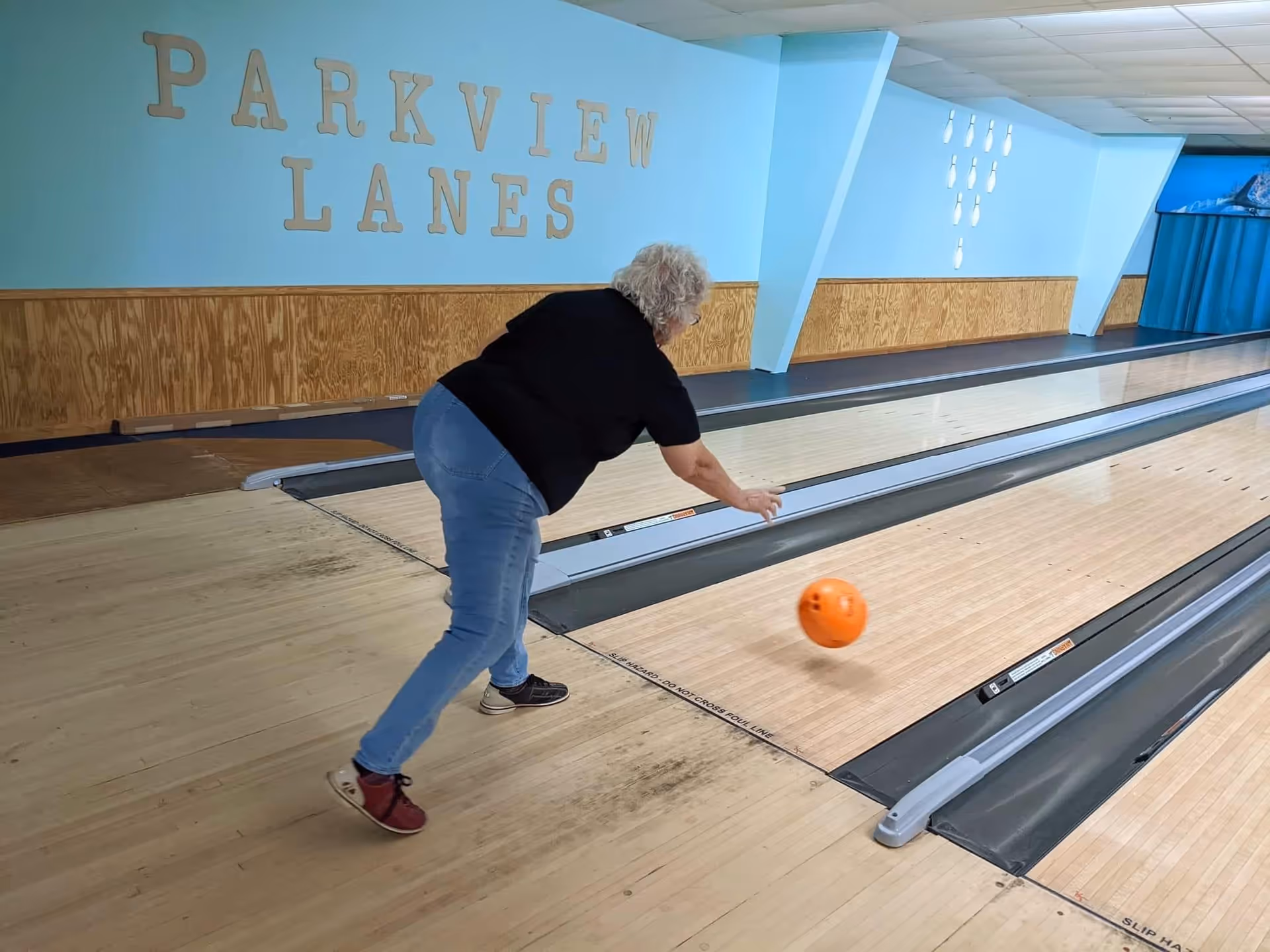 An elderly woman bowling at an indoor bowling alley named Parkview Lanes. She is releasing an orange bowling ball down the lane. The walls are painted light blue with wooden paneling along the lower half, and there are bowling pins displayed on the wall.