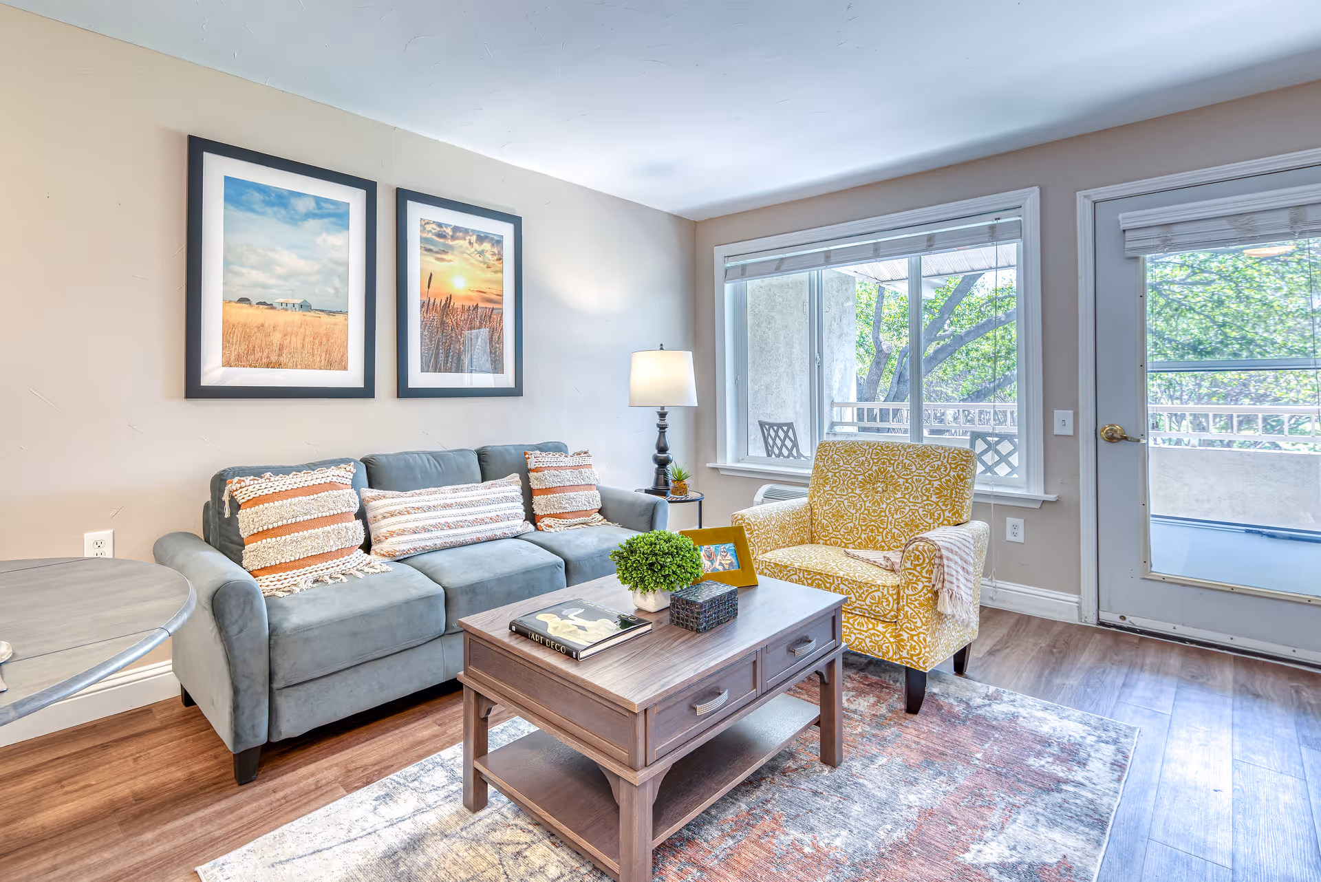 A bright living room with a gray sofa adorned with striped pillows, a yellow patterned armchair, a wooden coffee table with books and a small plant, a floor lamp, two framed landscape pictures on the wall, and large windows and a glass door letting in natural light and showing greenery outside.