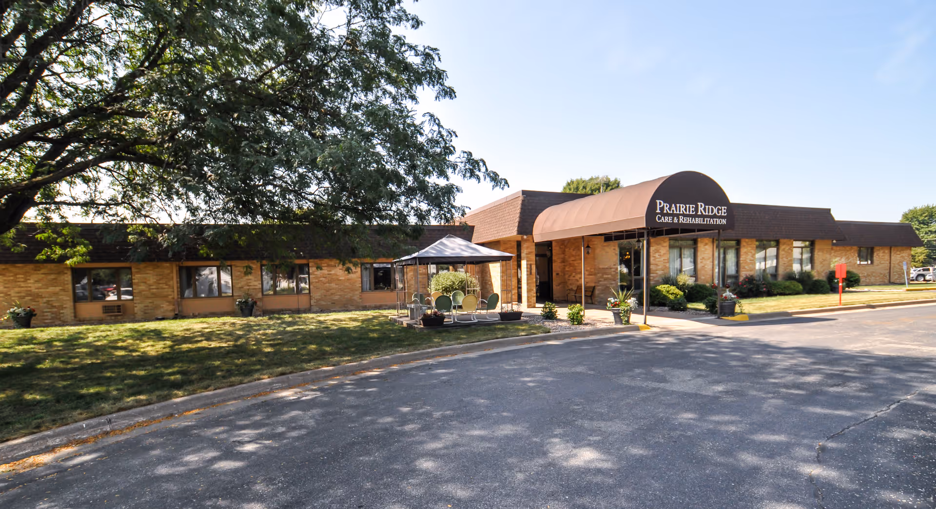 Exterior view of Prairie Ridge Care & Rehabilitation facility with a brown awning over the entrance, brick walls, a small outdoor seating area with a canopy, and a large tree providing shade on the lawn.