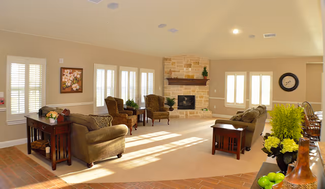 A spacious living room with beige walls and carpet, featuring two brown upholstered sofas, two patterned armchairs, a stone fireplace with a wooden mantle, multiple windows with white shutters, and wooden side tables. There are decorative plants and a painting on the wall, with sunlight streaming through the windows.