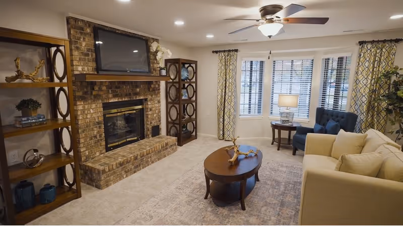 Cozy living room with a brick fireplace and mounted TV, sofa, coffee table, shelving units and a bay window.