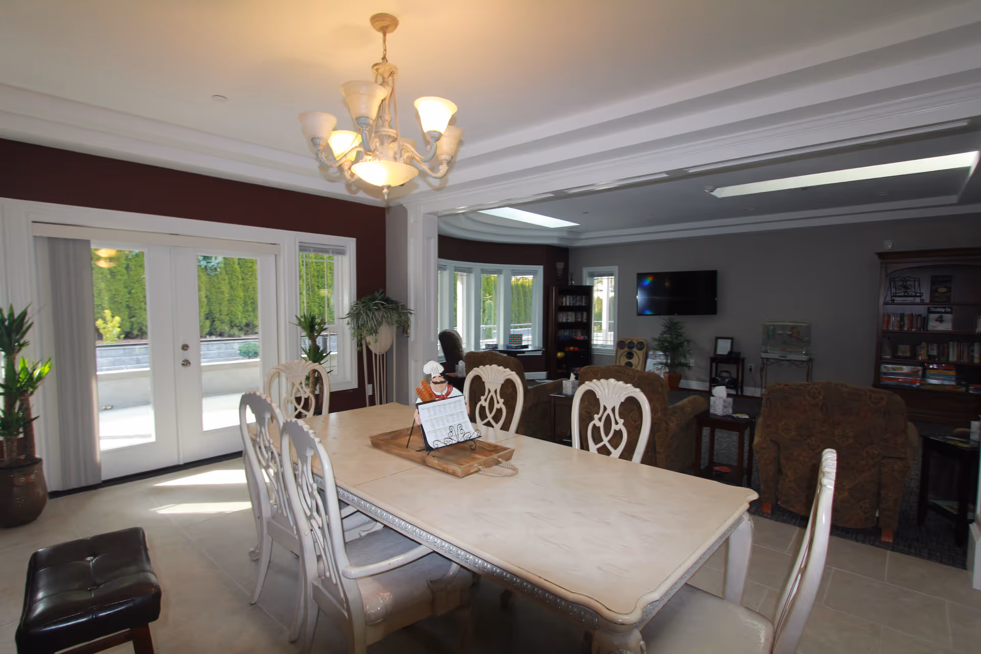 Interior view of a senior care home dining and living area featuring a long dining table with six chairs, a chandelier overhead, large windows and glass doors letting in natural light, and a living room with armchairs, a TV mounted on the wall, bookshelves, and plants.