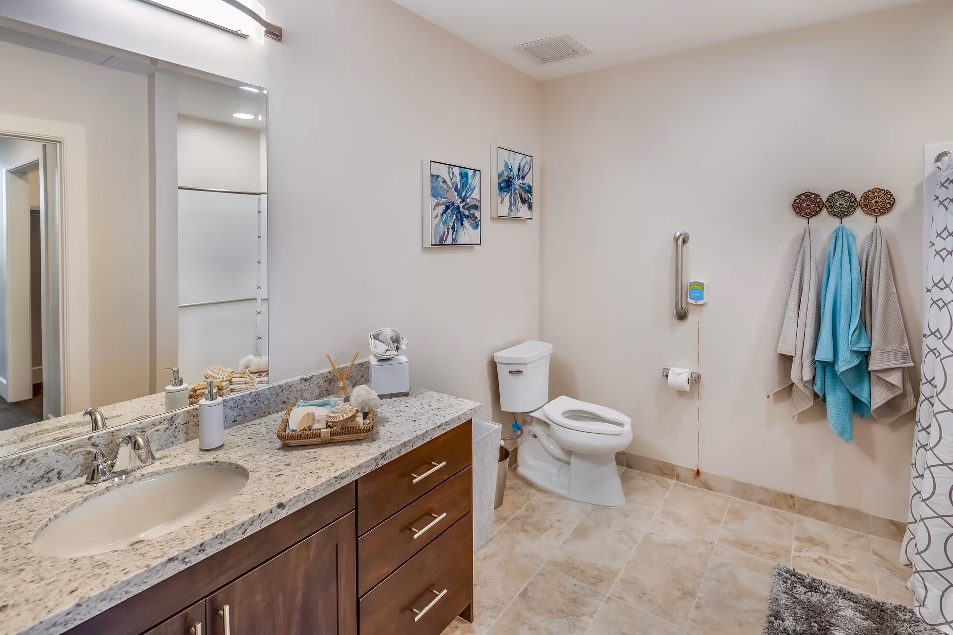 A clean and modern bathroom with a granite countertop vanity featuring a sink, soap dispenser, and decorative items. There is a toilet with a grab bar on the wall beside it, three towels hanging on decorative hooks, and a shower curtain partially visible on the right. The floor is tiled, and two abstract blue floral paintings hang on the wall above the toilet.