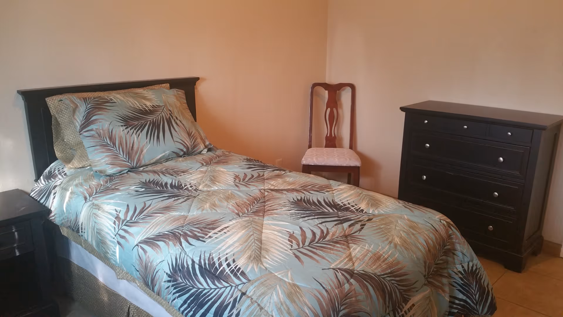 Single bed with tropical-patterned bedding, wooden headboard, nightstand, chair, and dark dresser in a simple bedroom.