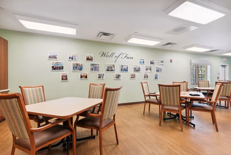 A bright and clean common area with several wooden tables and chairs arranged on a wood floor. On the light green wall, there is a display titled 'Wall of Fun' featuring multiple framed photos. The room has large ceiling lights and windows letting in natural light.