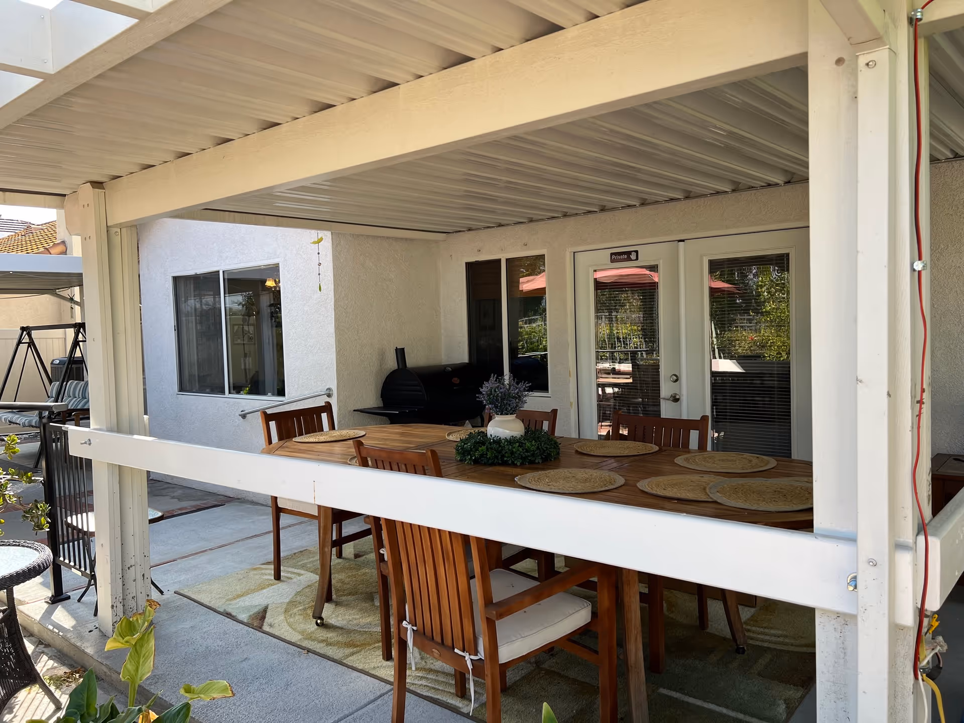Covered outdoor patio area with a wooden dining table and six chairs, each with a round placemat. A small plant centerpiece is on the table. The patio has a white roof and is adjacent to a white building with glass doors and windows. A black grill is visible in the corner, and there is a swing set in the background.