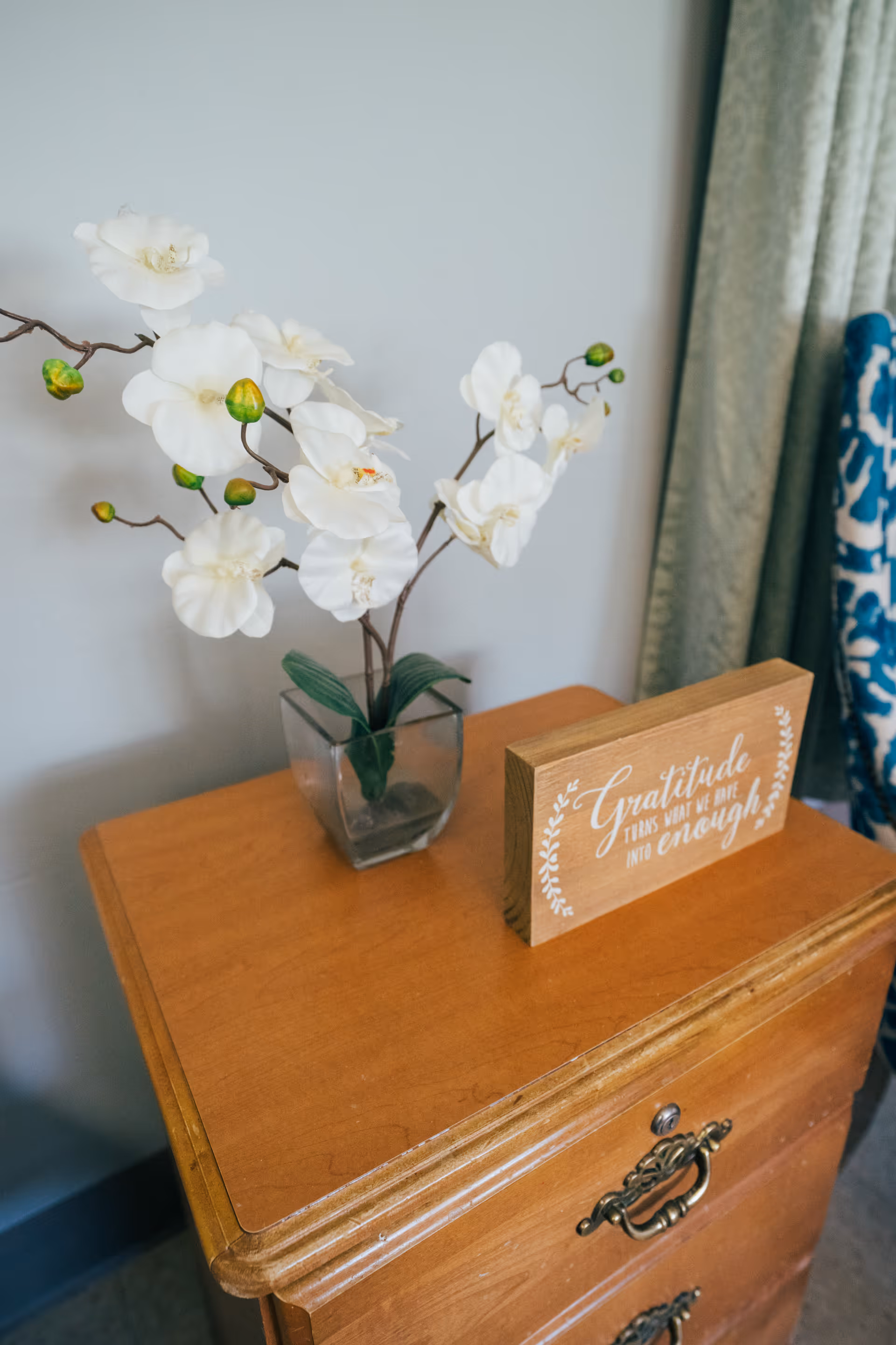 A wooden nightstand with a glass vase holding white orchid flowers and a wooden plaque with the inscription 'Gratitude turns what we have into enough.' A green curtain and part of a blue patterned chair are visible in the background.