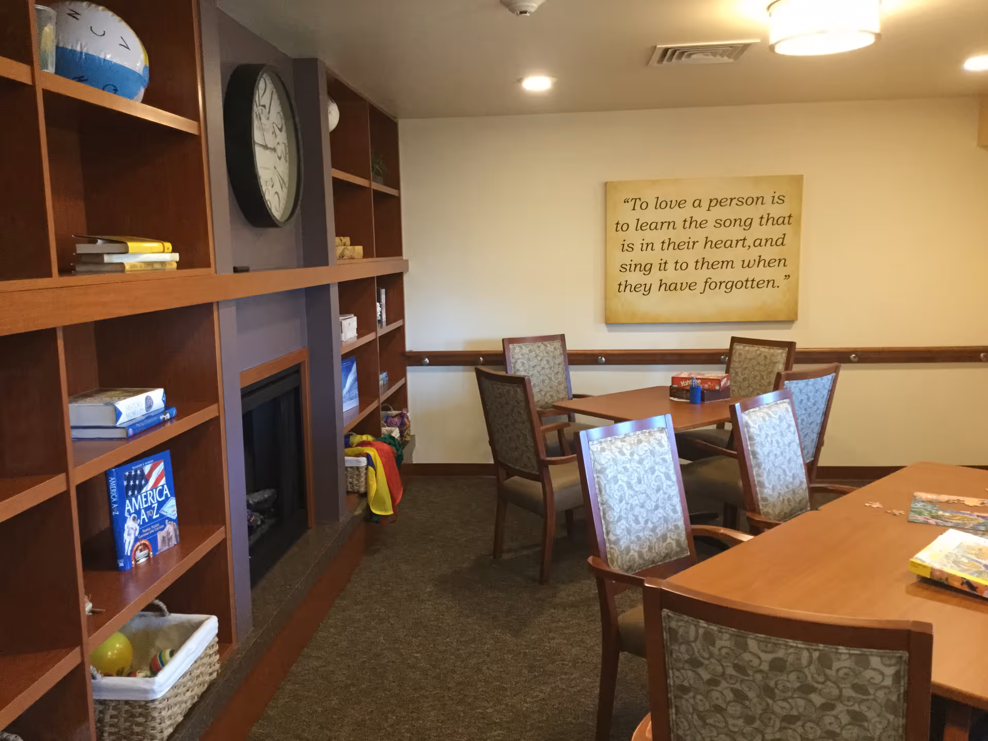 A cozy room with wooden bookshelves on the left filled with books, baskets, and a clock above a fireplace. There are two wooden tables with patterned cushioned chairs around them. On the wall, there is a framed quote that reads, 'To love a person is to learn the song that is in their heart, and sing it to them when they have forgotten.' The room has a carpeted floor and soft lighting from ceiling fixtures.