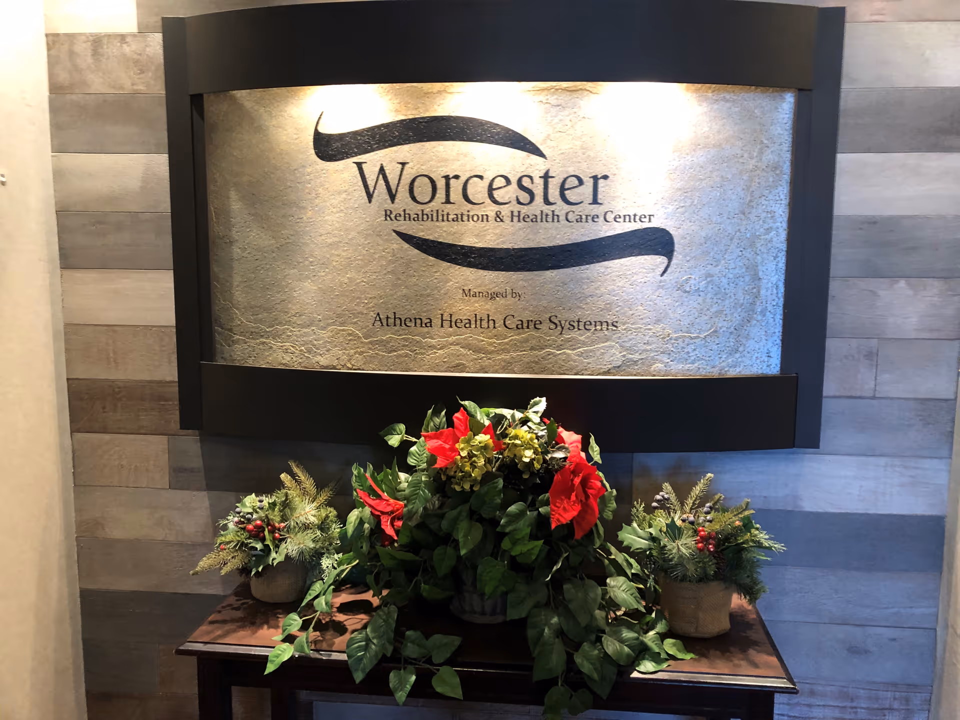 A decorative sign for Worcester Rehabilitation & Health Care Center managed by Athena Health Care Systems mounted on a textured wall above a wooden table with three floral arrangements featuring green leaves and red flowers.