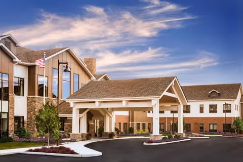 Exterior view of a senior living facility named Montage Mason featuring a covered entrance with white pillars, a paved driveway, landscaped greenery, and an American flag on a flagpole under a partly cloudy sky.