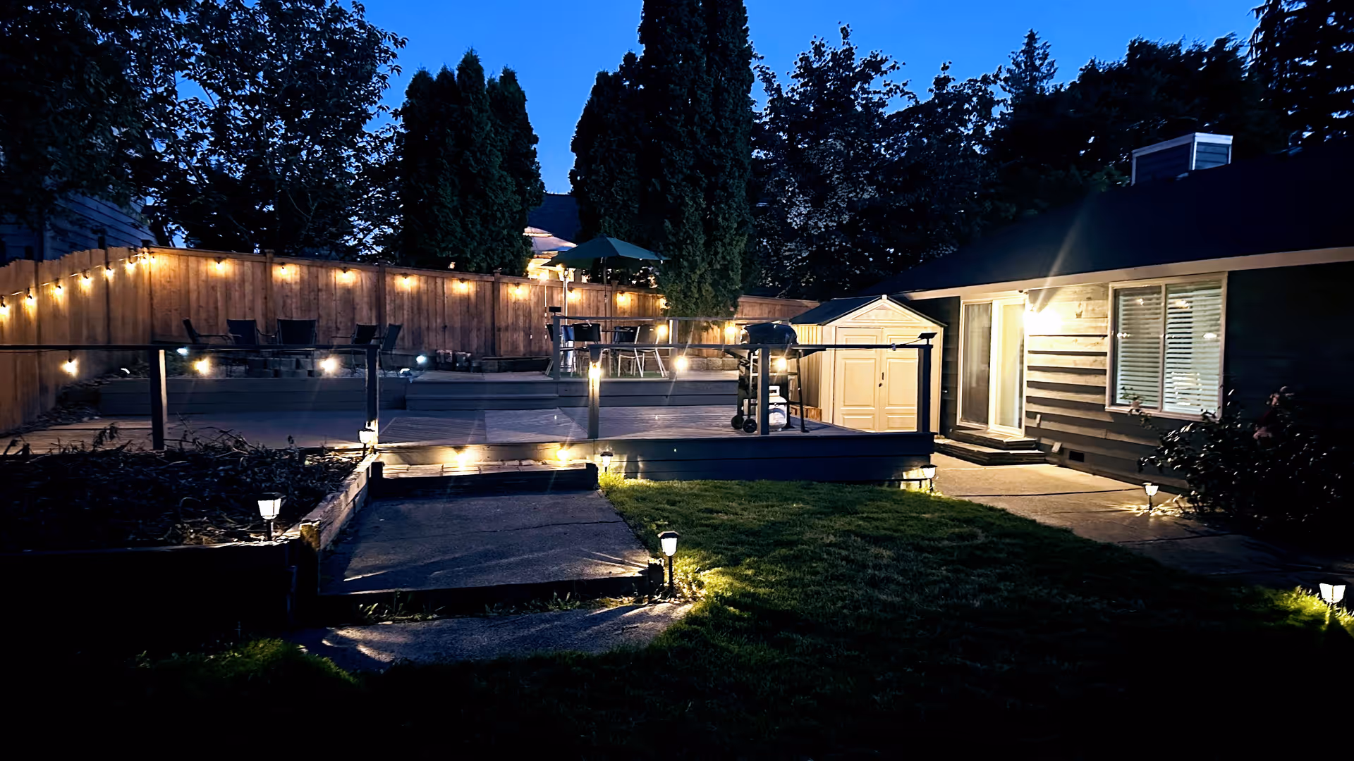 Outdoor backyard area at dusk with a wooden deck decorated with string lights, patio furniture, a barbecue grill, a small shed, and a house with illuminated windows and door. The area is surrounded by a wooden fence and tall trees.