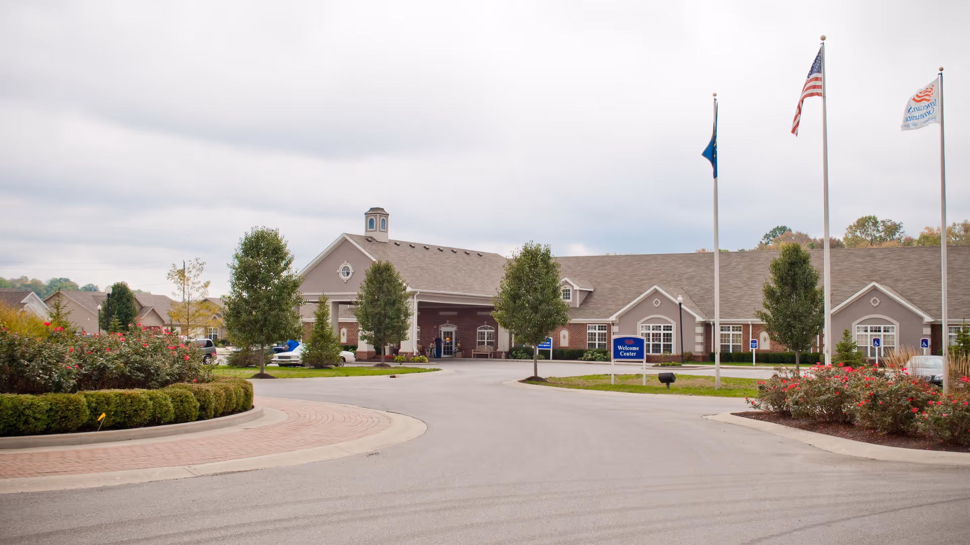 Exterior view of Meadow Lakes Assisted Living and Garden Homes facility showing a large building with a covered entrance, surrounded by neatly trimmed bushes, trees, and flower beds. Three flagpoles with flags are visible near the entrance, along with a blue sign that reads 'Welcome Center'. The sky is overcast.