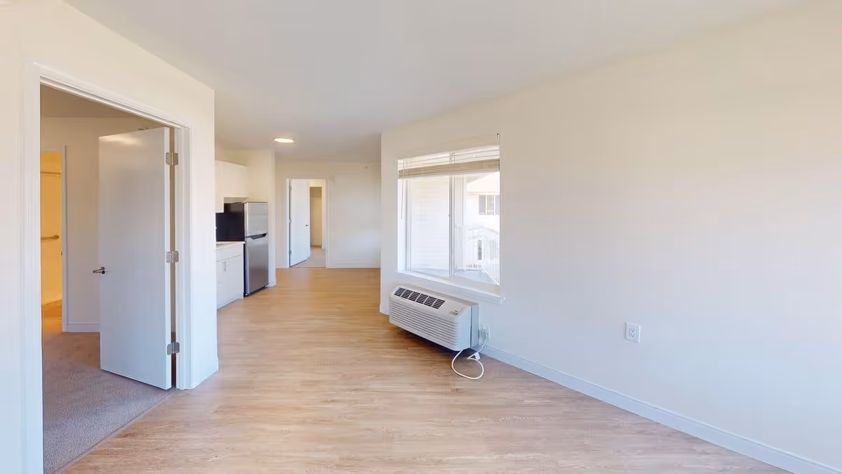 Interior view of an empty apartment unit with light wood flooring and white walls. The image shows a small kitchen area with a refrigerator and cabinets on the left side, an open doorway leading to another room, a large window with blinds, and a wall-mounted air conditioning unit below the window.