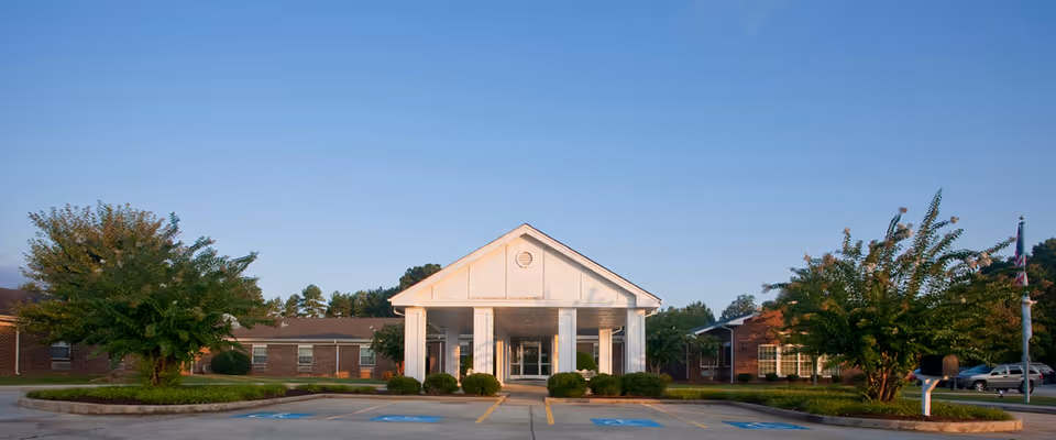 Front exterior view of The Laurels of Salisbury facility with a white entrance canopy supported by columns, surrounded by trees and a parking lot with handicap spaces.