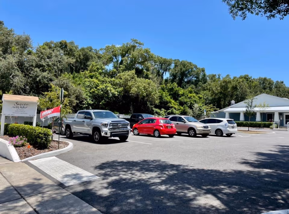 Parking lot of Sweet Water of Orlando facility with several parked cars including a silver pickup truck, a red compact car, and other vehicles. The facility building is visible on the right side with a white exterior and green shutters. Trees and greenery surround the area under a clear blue sky.