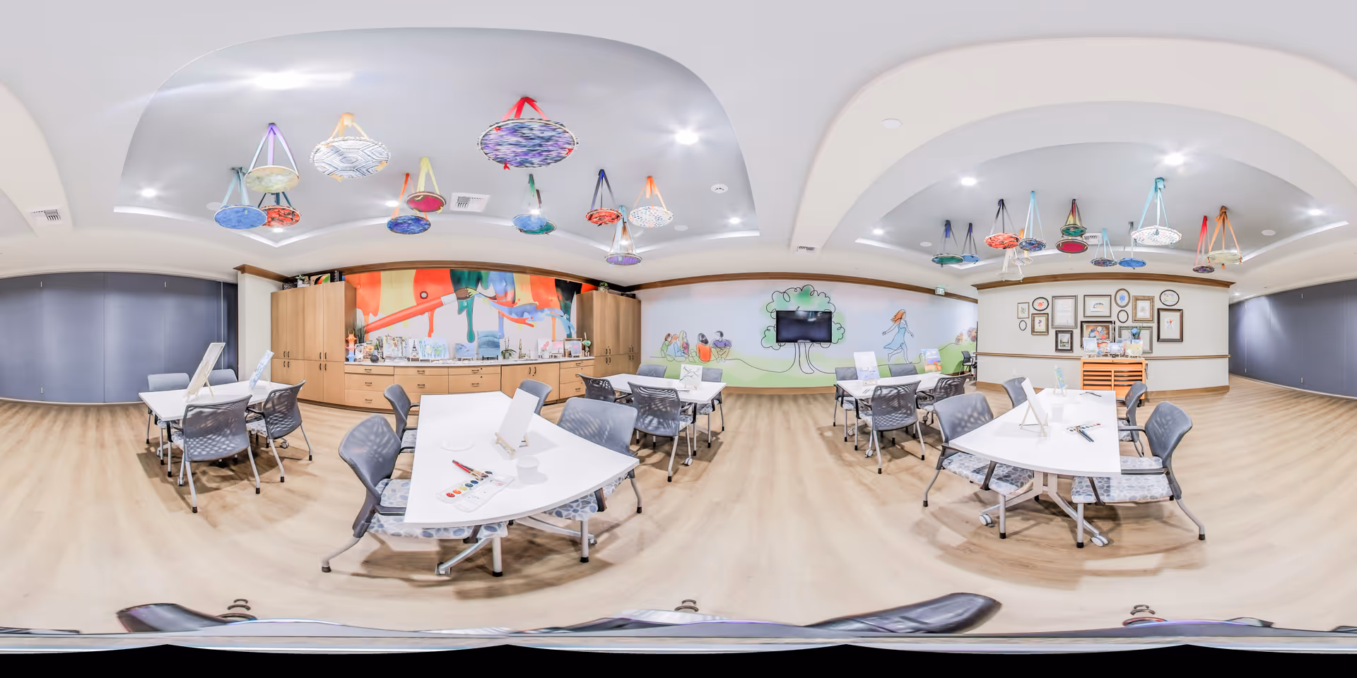 A spacious, well-lit activity room with several tables and chairs arranged for group activities. The ceiling features colorful hanging decorations. The walls have playful murals and a mounted TV. Cabinets and shelves along the back wall hold various supplies and materials.