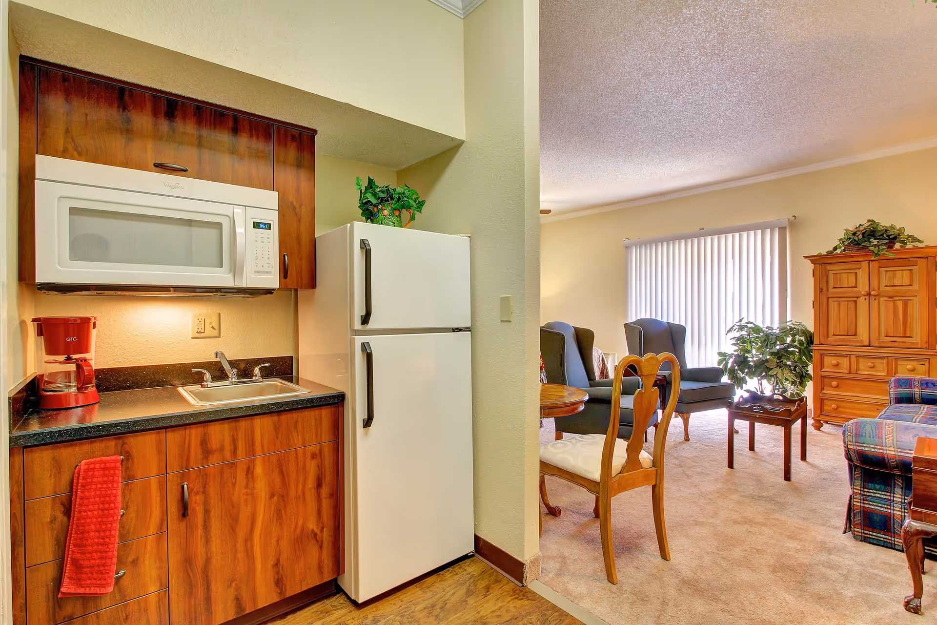 A small kitchen area with wooden cabinets, a white microwave, a white refrigerator, a red coffee maker, and a sink with a red towel hanging on the cabinet. Adjacent to the kitchen is a living room with two blue armchairs, a wooden dining chair, a wooden cabinet, a small wooden coffee table, a plaid sofa, and a large window with vertical blinds.